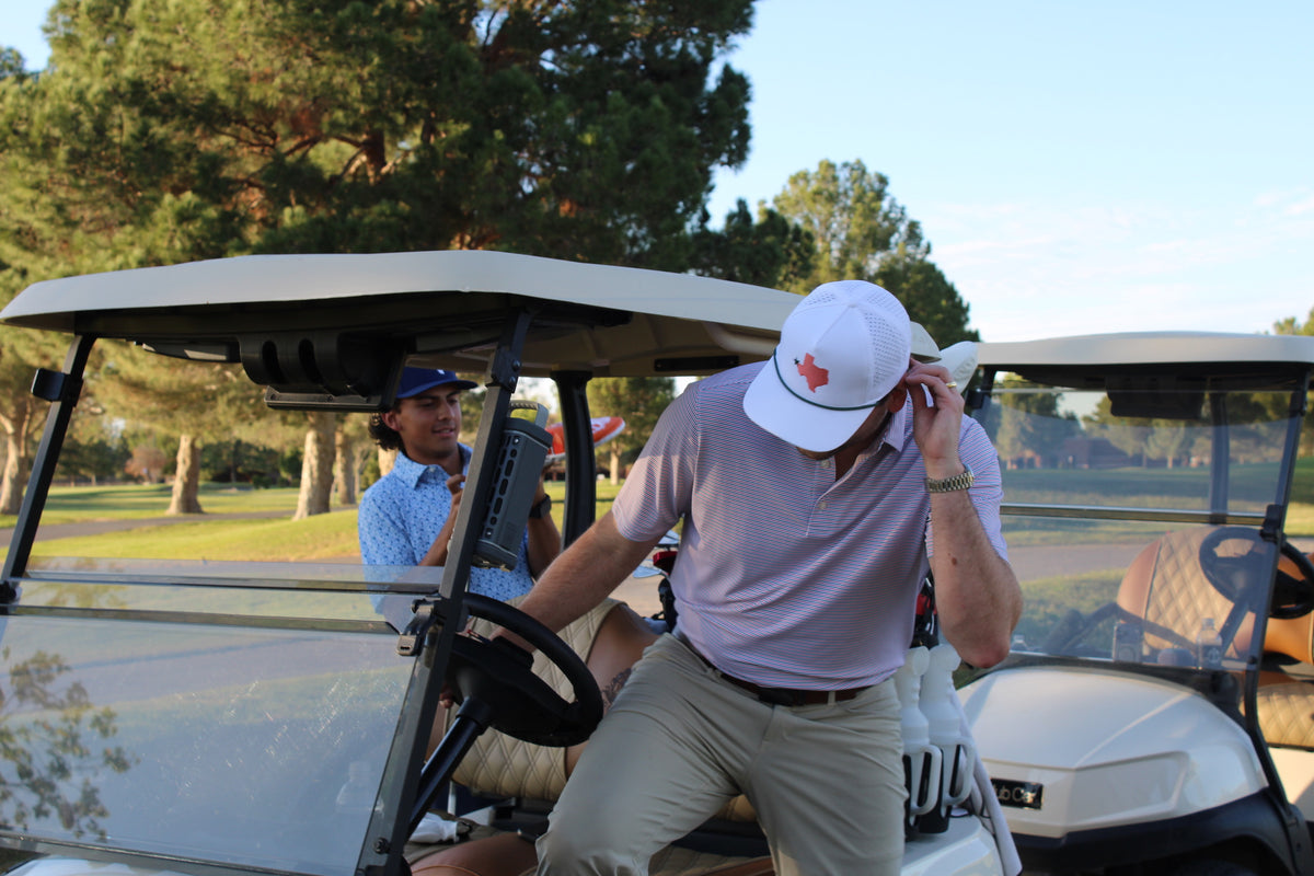 A man in a golf cart on a golf course, wearing a white polo shirt, khaki pants, and a white cap with a red logo. Another person is seated inside the cart in the background.