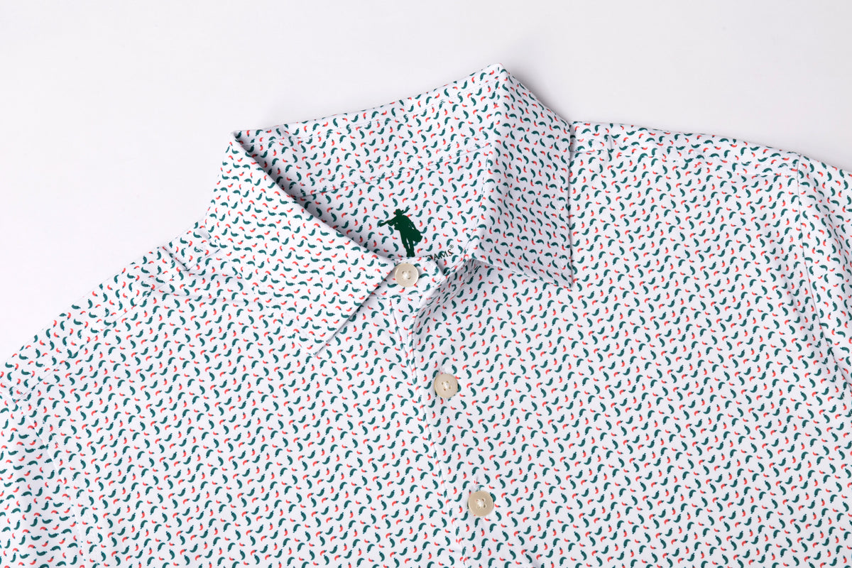 A close-up of a white button-down shirt with a dense, small-scale multicolor print of red, blue, and green motifs, featuring a classic collar and a small green embroidered polo player logo on the inner neck label.