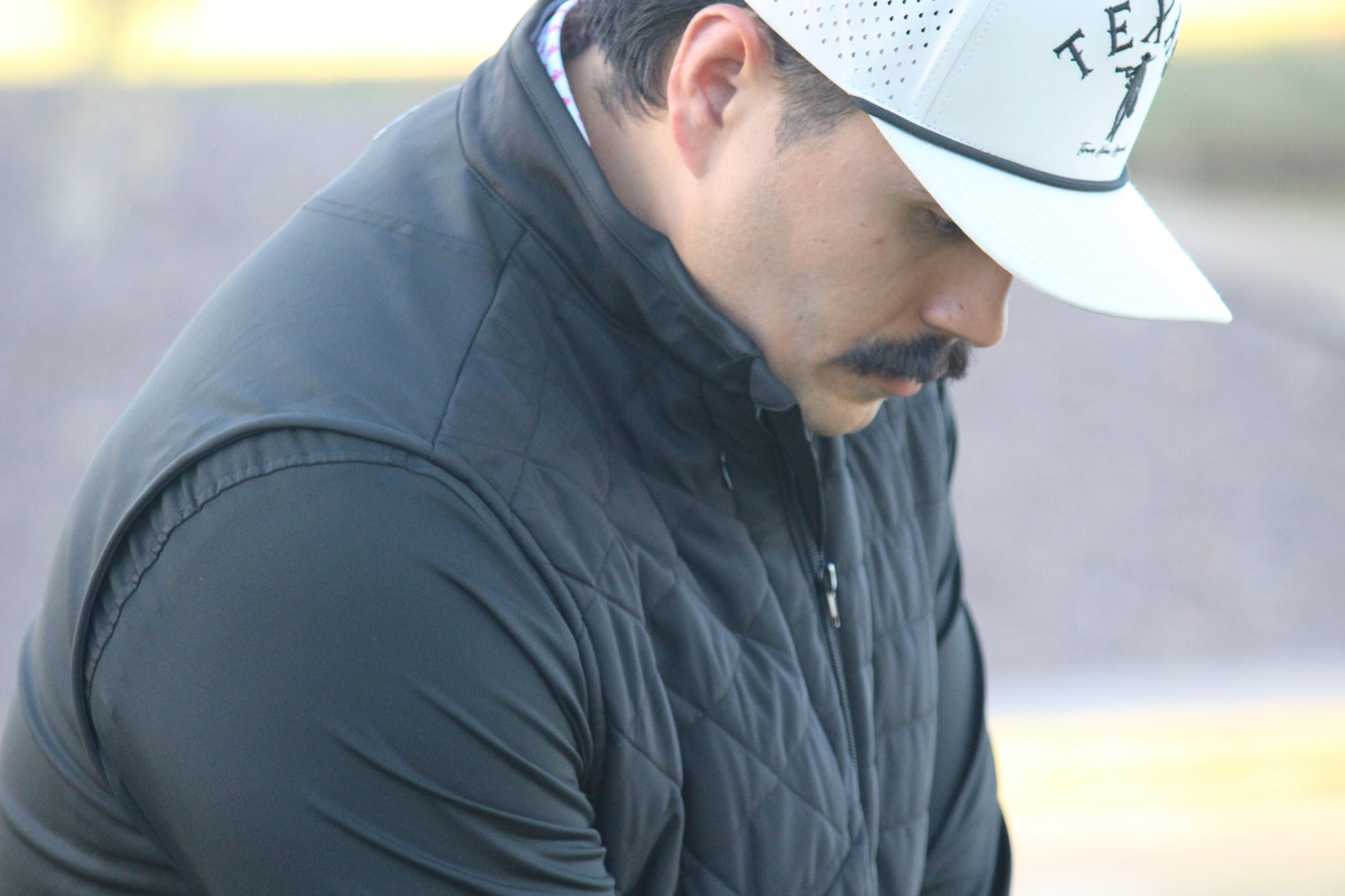 Man wearing a black quilted jacket and white cap with a logo, looking down.