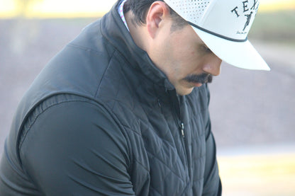 Man wearing a black quilted jacket and white cap with a logo, looking down.