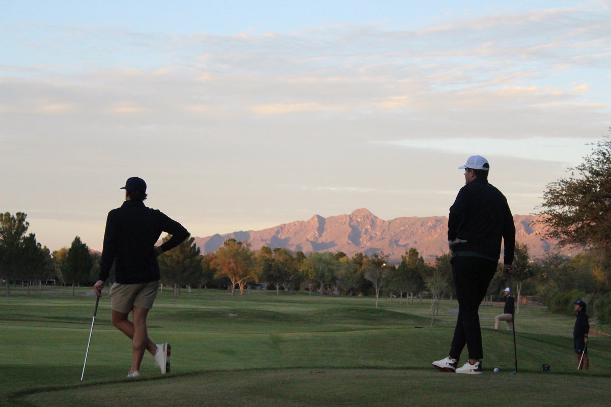 Two golfers on a golf course at sunset with mountains in the background. One golfer on the left wears a black long-sleeve top, khaki shorts, and a black cap while holding a golf club.