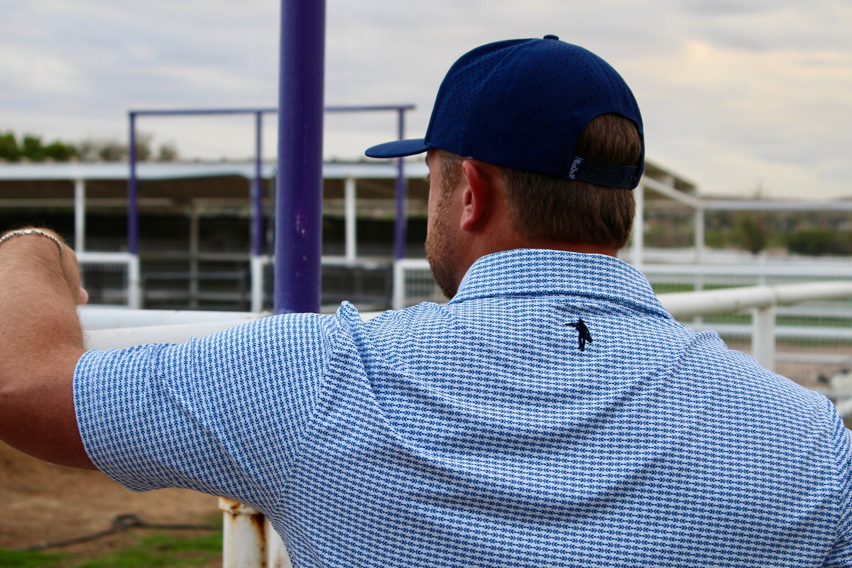 A man viewed from behind wearing a blue and white checkered polo shirt and a navy blue baseball cap, standing at an outdoor racetrack.