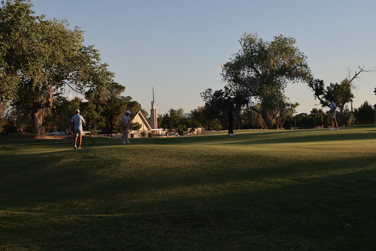 A wide outdoor lawn scene at sunset with several people standing or walking across the grass, large trees on both sides, and a small building in the distance under a pale blue sky.